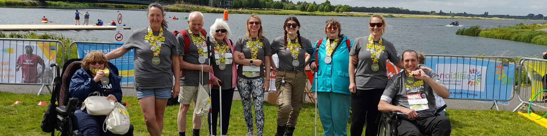 group photo of the Coalition Superhero Tri participants at the event in front of a large lake. All of the participants are wearing a large yellow and silver medal.