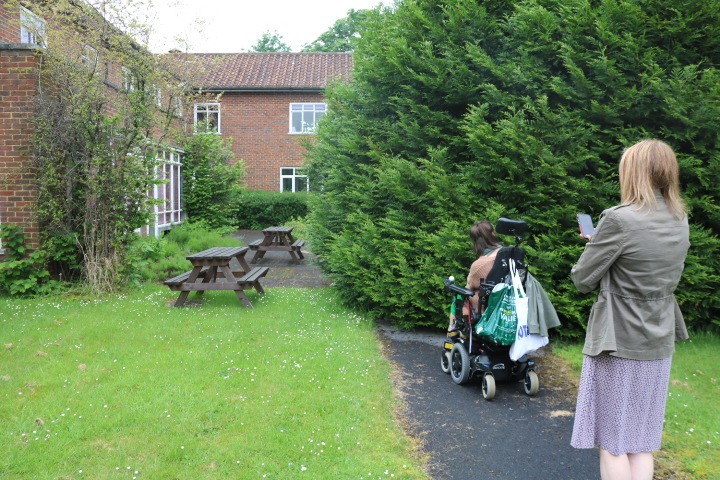 The Coalition's CEO, Nikki, and our work experience student, Is, are walking and wheeling on a path to picnic benches at the Astolat office that is blocked by a large overgrown tree.