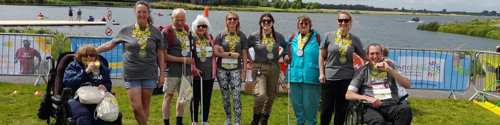 Group photo of the Coalition Superhero Tri participants at the event in 2023 in front of a large lake. All of the participants are wearing a large yellow and silver medal.