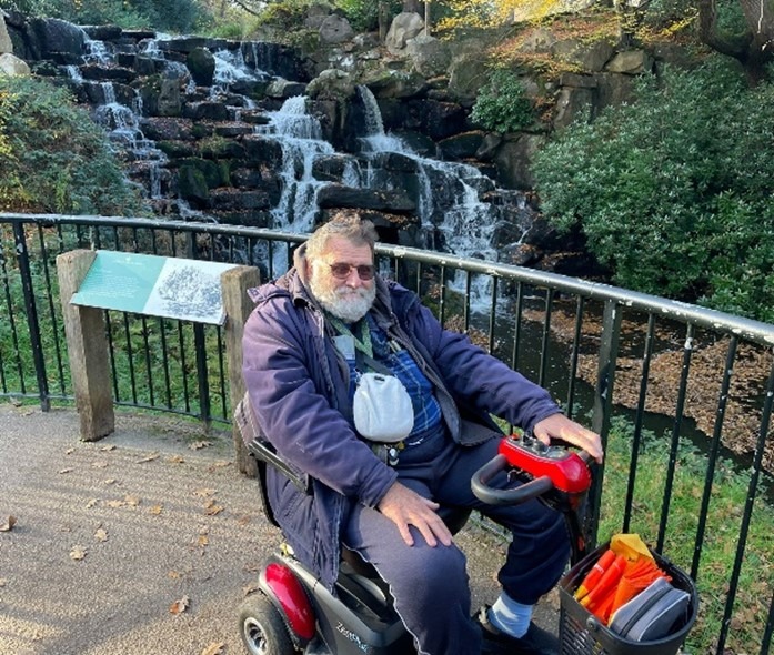 David M on his mobility scooter in front of a waterfall at Windsor Great Park. 