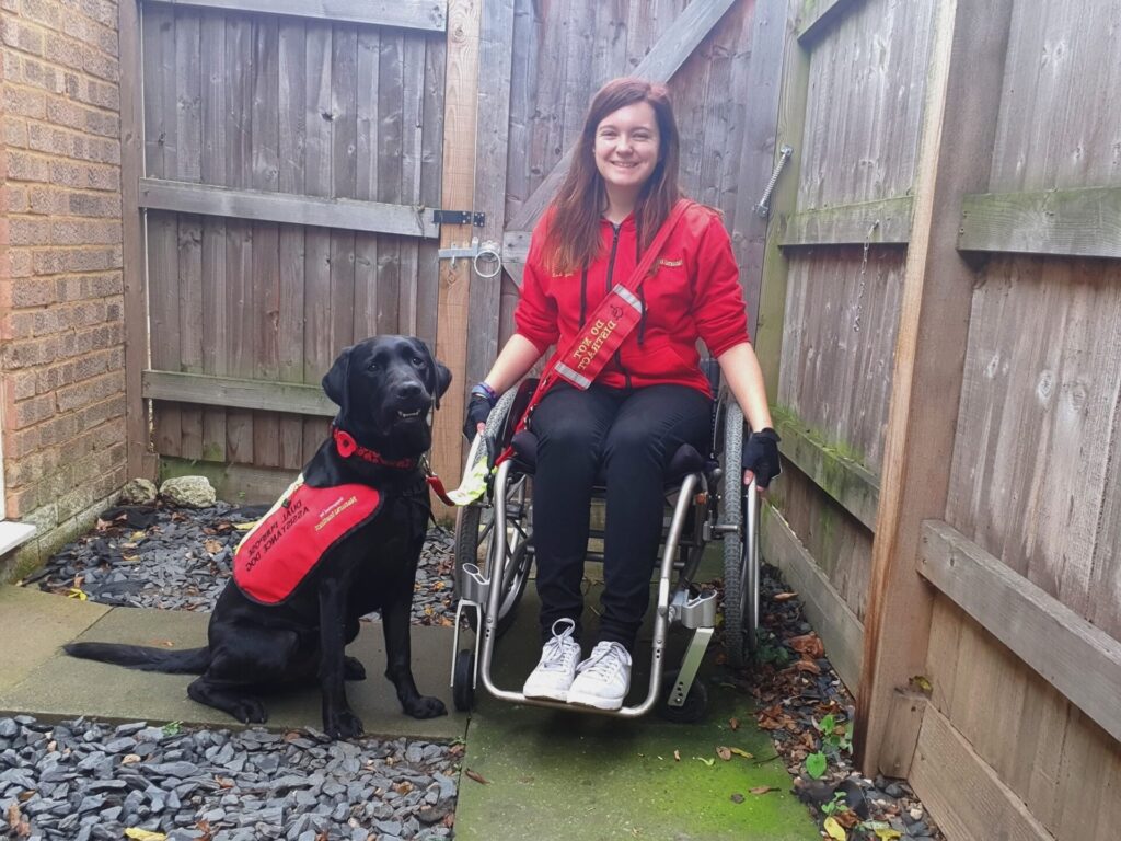 Megan is in a wheelchair with Rowley her assistance dog next to her, they are outside.