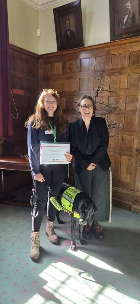 Megan smiling with her assistance dog Rowley, she's holding a certificate and is next to the Chief Executive of Reigate and Banstead Borough Council, Mari Roberts-Wood.